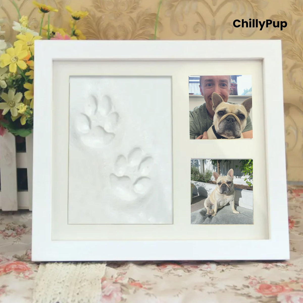 White photo frame with paw print and two pictures of a dog, on a floral surface with flowers in the background.